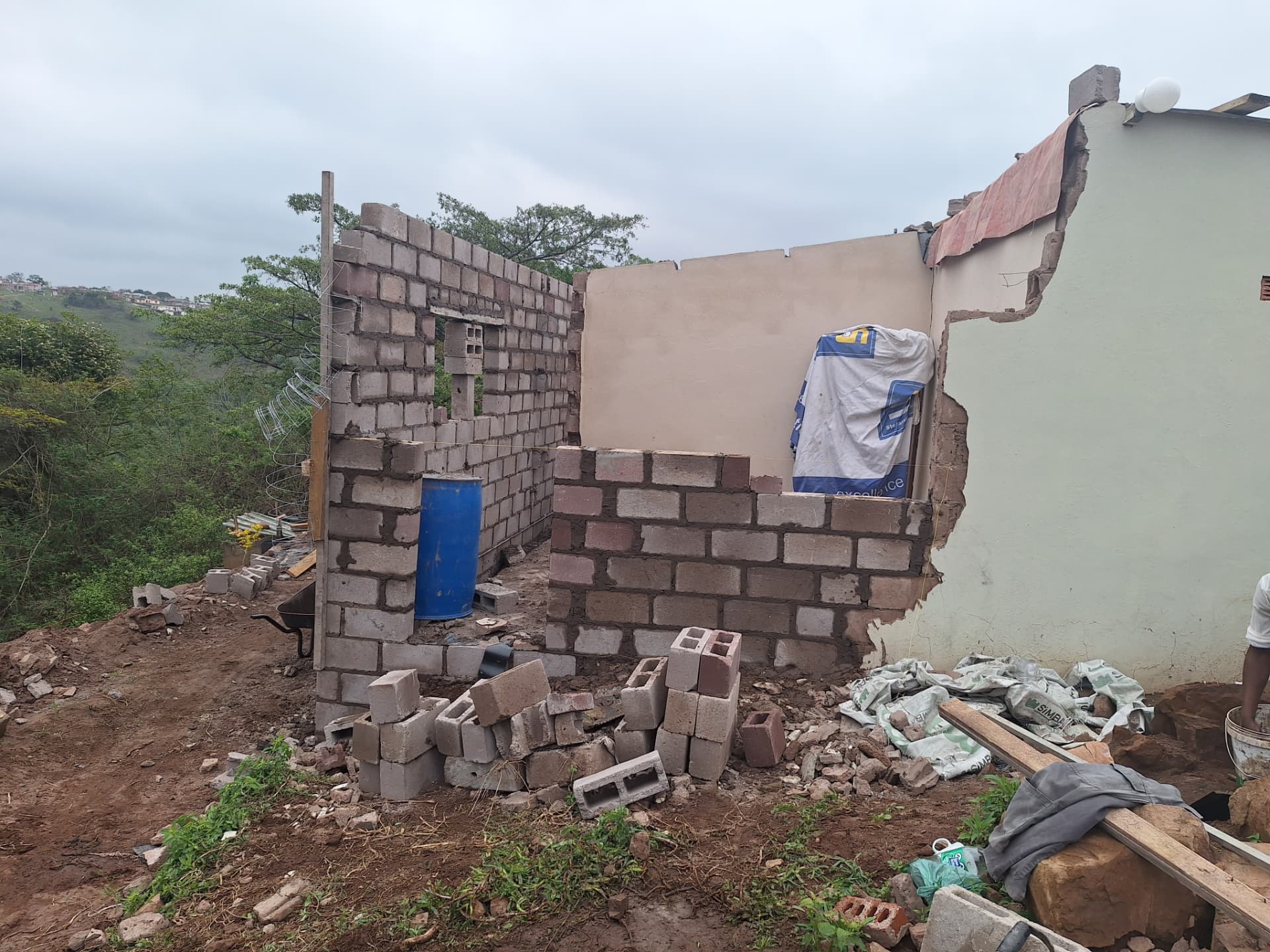 Interior view of the unfinished block walls joining the old green wall, with blocks and a wheelbarrow on the dirt floor
